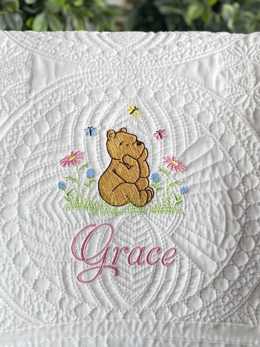 White quilt featuring an embroidered Classic Pooh Bear surrounded by colorful butterflies and flowers, with the custom name Grace stitched below on the Personalized Classic Pooh Bear Meadow & Butterfly Baby Quilt | Custom Name Heirloom Blanket.