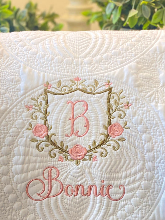 Close-up of a white Personalized Embroidered Rose Crest Monogram Baby Quilt | Heirloom Keepsake featuring a pink embroidered letter B inside a rose and leaf wreath with the name Bonnie stitched below.
