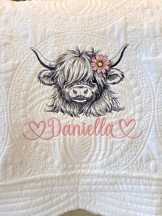 Close-up of a personalized embroidered highland cow with a pink daisy and the name Daniella in pink script on a white quilt featuring detailed stitching, showcasing the Personalized Embroidered Highland Cow Daisy Baby Quilt | Custom Name Heirloom Blanket.