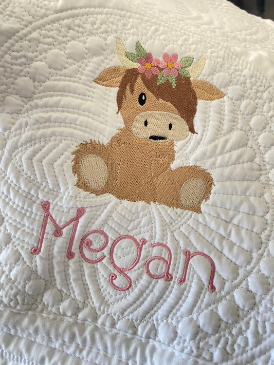 Personalized Embroidered Highland Cow Flower Crown Baby Quilt | Custom Name Heirloom Blanket featuring a detailed brown highland cow with a pink flower crown and the name Megan embroidered in pink on a textured white quilt.