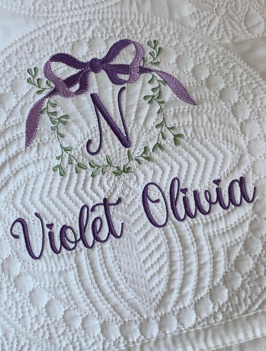 Personalized Vine & Bow Monogram Baby Quilt | Custom Embroidered Heirloom Blanket featuring a purple monogram N with a vine and bow design, and the name Violet Olivia embroidered on white quilted fabric.