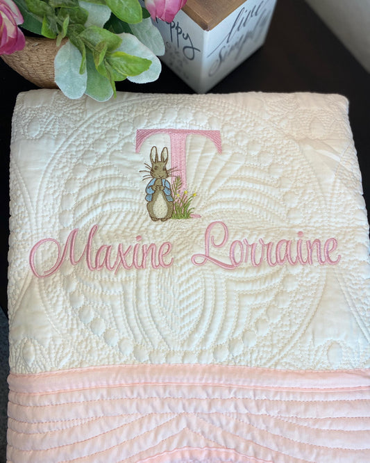 Folded Personalized Embroidered Bunny Baby Quilt | Classic Storybook Heirloom Keepsake | Custom Name Blanket with pink stitched name Maxine Lorraine and embroidered bunny monogram on white quilted fabric.