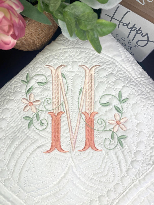 Close-up of a white quilt embroidered with an elegant floral interlocking monogram "M," showcasing delicate pink and green flower and vine details on the Personalized Elegant Floral Interlocking Monogram Baby Quilt | Custom Embroidered Heirloom Blanket.