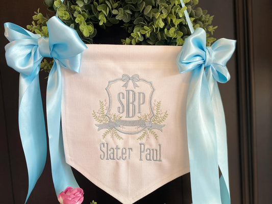 Personalized Embroidered Baby Birth Announcement Banner | Hospital Door & Bassinet Sign with light blue satin ribbons, custom monogram, and name Slater Paul on white fabric hanging against greenery.