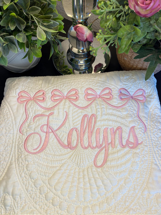 Personalized Pink Bow Baby Quilt | Custom Name Heirloom Blanket for Girls with delicate pink embroidered bows and custom name on a soft white quilt, surrounded by greenery.