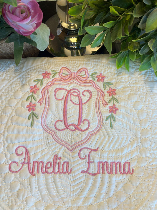 Personalized Floral Wreath & Bow Monogram Baby Quilt | Custom Name Heirloom Keepsake featuring delicate pink embroidered monogram Q with floral wreath and custom name Amelia Emma on a white quilted fabric.