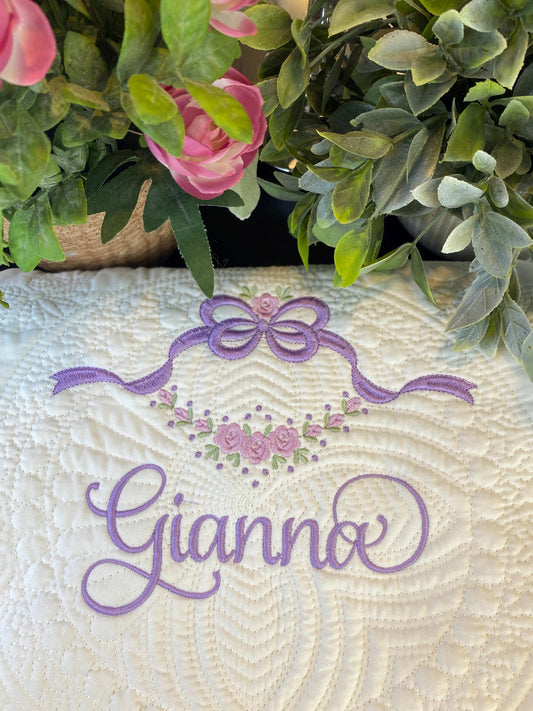 Close-up of a Personalized Floral Vine & Bow Baby Quilt | Custom Name Heirloom Blanket featuring delicate lavender embroidery of a bow and floral vine with the name Gianno on a textured white quilt surrounded by greenery and pink flowers.