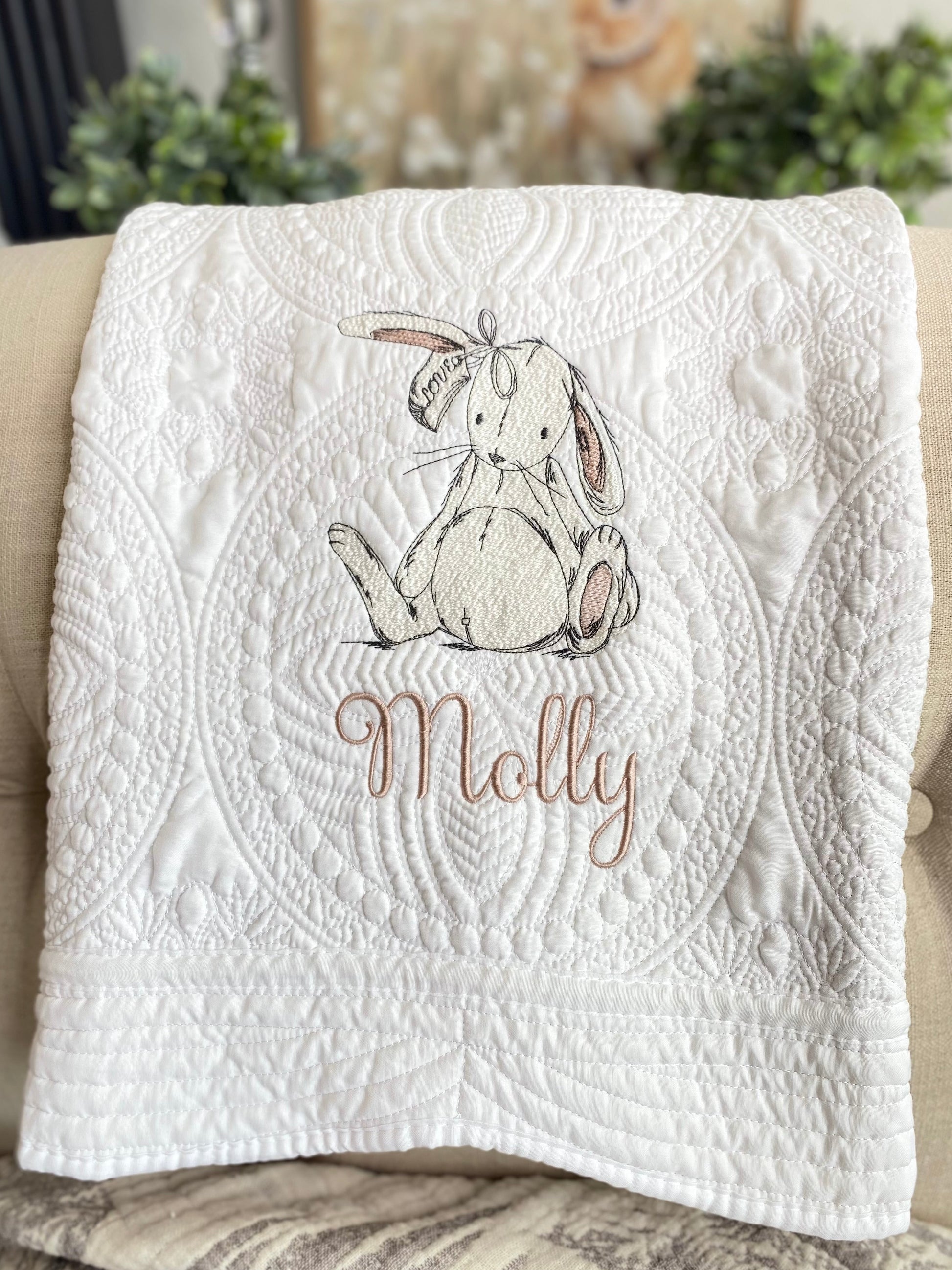 White quilt with intricate stitched patterns featuring a delicate embroidered sketch of a bunny rabbit and the custom name Molly, showcasing the Personalized Sketch Bunny Rabbit Baby Quilt | Custom Name Heirloom Keepsake Blanket.