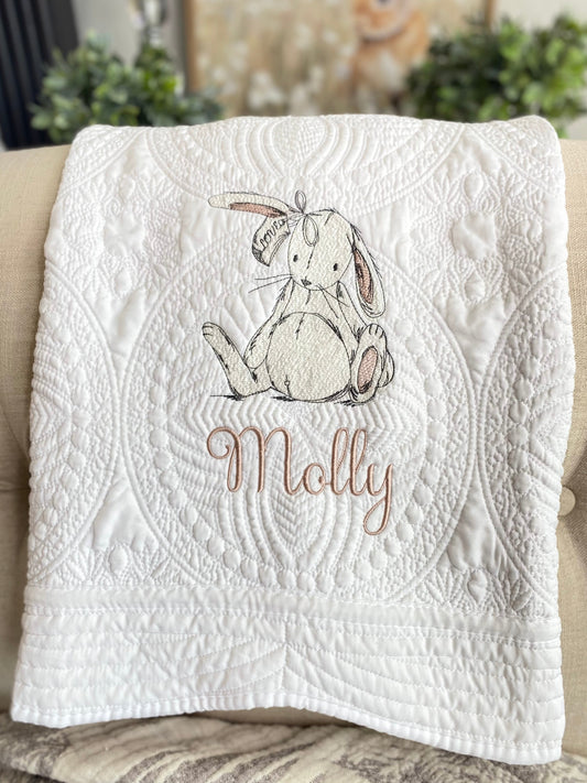 White quilt with intricate stitched patterns featuring a delicate embroidered sketch of a bunny rabbit and the custom name Molly, showcasing the Personalized Sketch Bunny Rabbit Baby Quilt | Custom Name Heirloom Keepsake Blanket.
