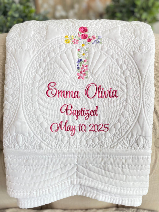 White quilt with detailed floral cross embroidery and pink script personalization reading Emma Olivia Baptized May 10 2025, showcasing personalized garden floral cross baptism baby quilt | custom name & date christening heirloom.
