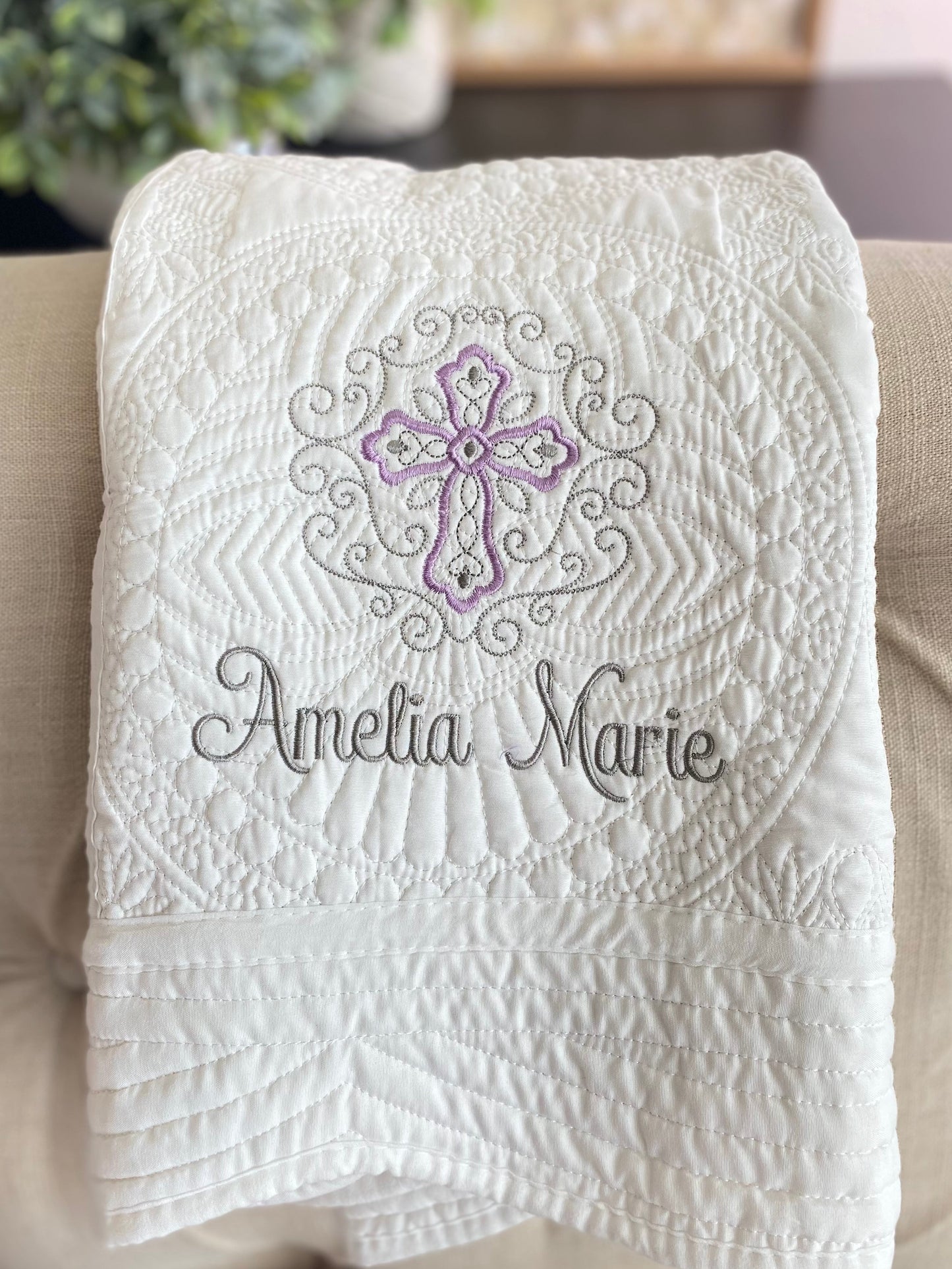 White quilt with intricate embossed filigree design featuring a lavender and gray cross and custom name embroidery, showcasing the Personalized Lavender Filigree Cross Baby Quilt | Custom Name Christening Heirloom Blanket for Girls.