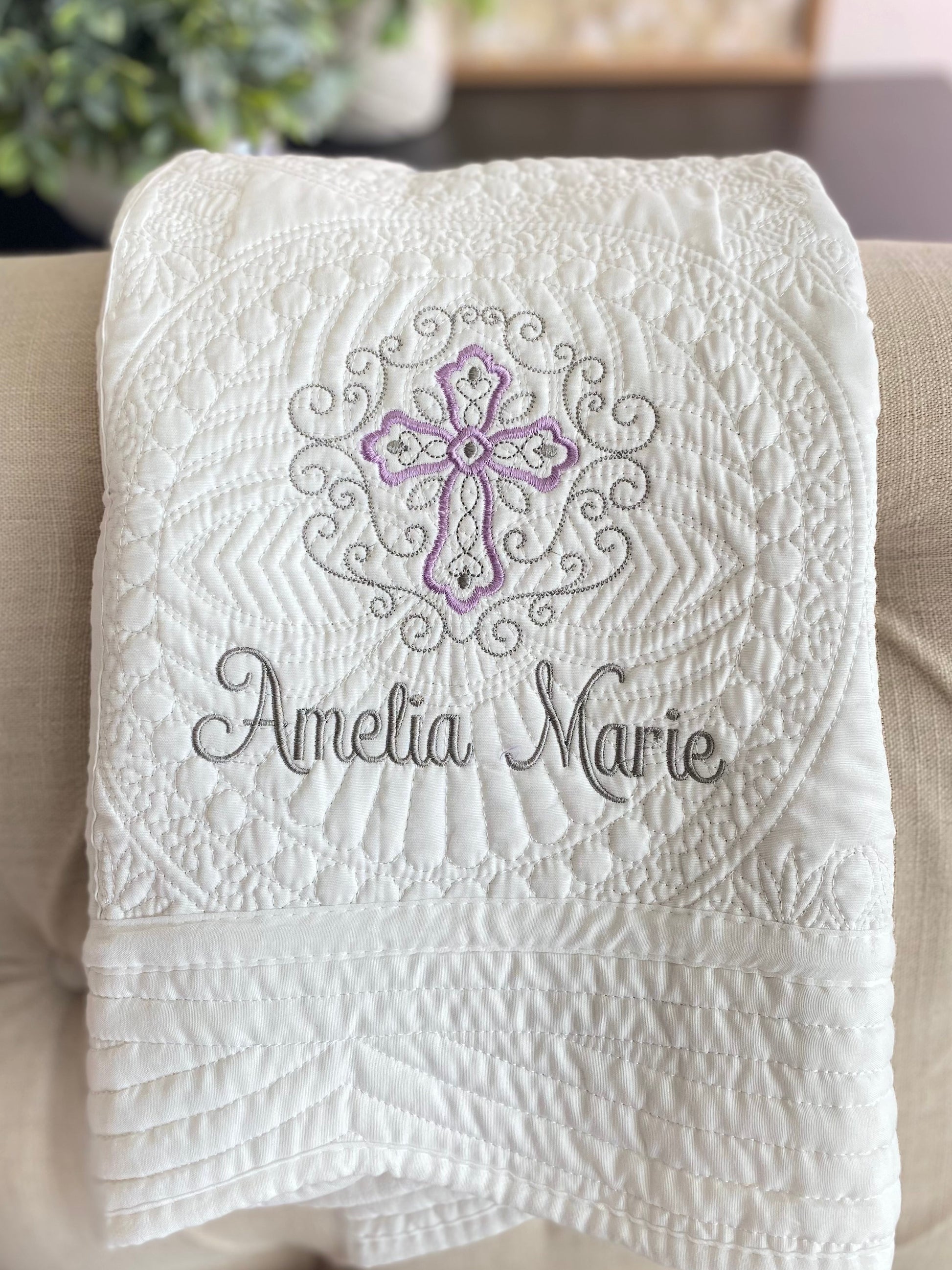 White quilt with intricate embossed filigree design featuring a lavender and gray cross and custom name embroidery, showcasing the Personalized Lavender Filigree Cross Baby Quilt | Custom Name Christening Heirloom Blanket for Girls.