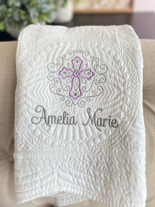 White quilt with intricate embossed filigree design featuring a lavender and gray cross and custom name embroidery, showcasing the Personalized Lavender Filigree Cross Baby Quilt | Custom Name Christening Heirloom Blanket for Girls.