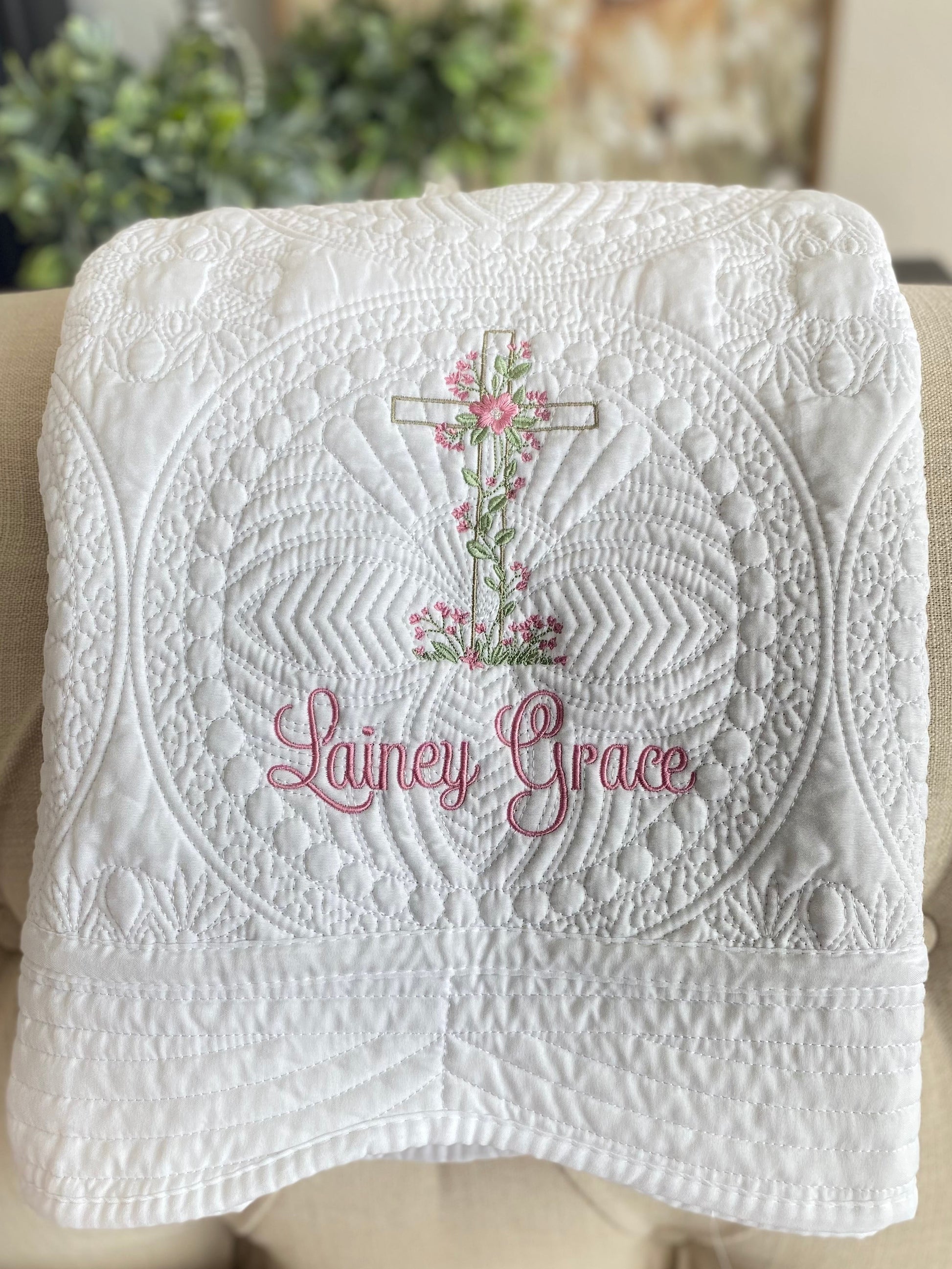 White quilt featuring intricate stitching, embroidered floral heart and vine cross, and the name Lainey Grace in pink script, personalized Heart & Vine Cross Baptism Baby Quilt | Custom Name Christening Heirloom Blanket for Girls.