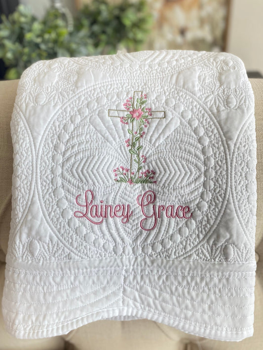 White quilt featuring intricate stitching, embroidered floral heart and vine cross, and the name Lainey Grace in pink script, personalized Heart & Vine Cross Baptism Baby Quilt | Custom Name Christening Heirloom Blanket for Girls.