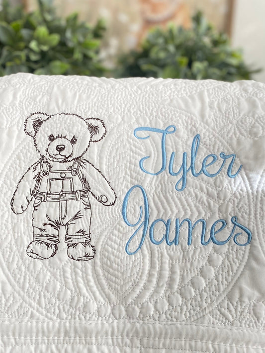 Close-up of a Personalized Embroidered Teddy Bear in Overalls Baby Quilt | Custom Name Heirloom Blanket featuring a detailed teddy bear in stitched overalls and the name Tyler James in blue embroidery on a white quilted background.