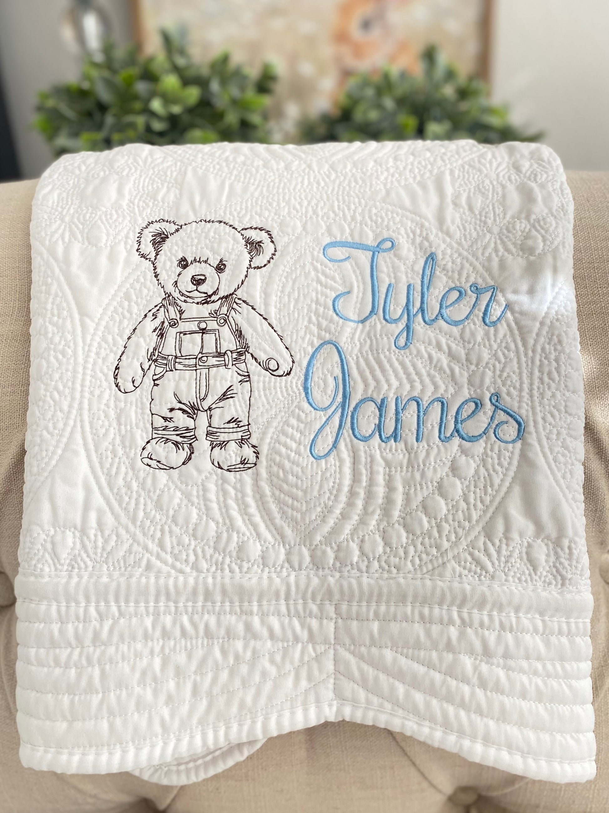 Personalized Embroidered Teddy Bear in Overalls Baby Quilt | Custom Name Heirloom Blanket featuring a detailed teddy bear in overalls and the name Tyler James embroidered in light blue on a white quilted background.