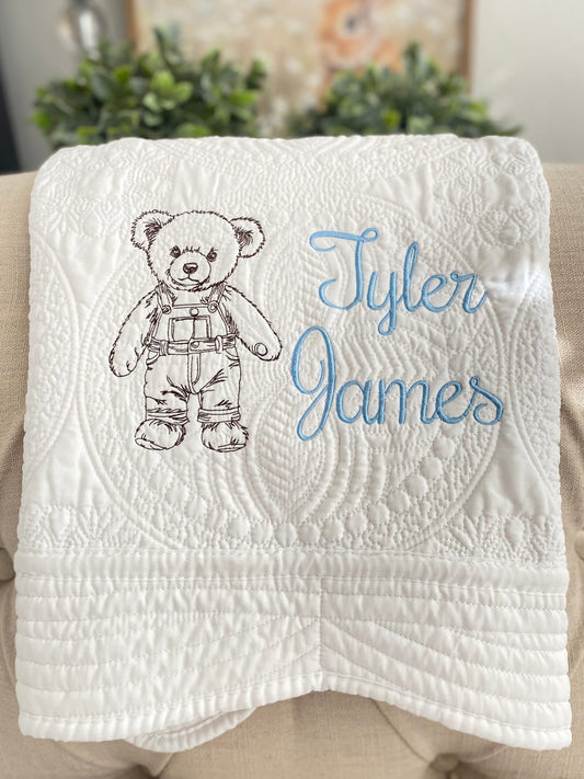 Personalized Embroidered Teddy Bear in Overalls Baby Quilt | Custom Name Heirloom Blanket featuring a detailed teddy bear in overalls and the name Tyler James embroidered in light blue on a white quilted background.
