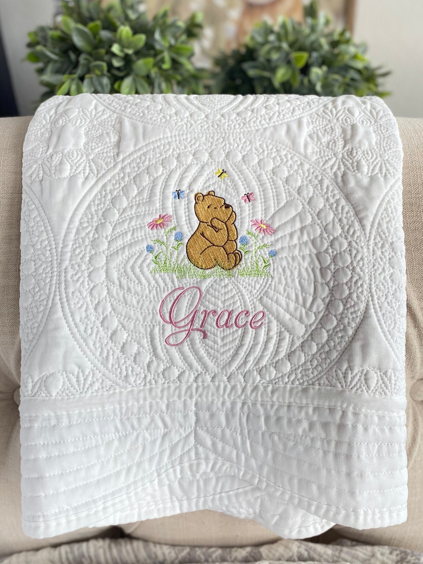 White quilt with intricate quilting patterns featuring an embroidered Classic Pooh Bear sitting in a meadow with butterflies and flowers, personalized with the name Grace, part of the Personalized Classic Pooh Bear Meadow & Butterfly Baby Quilt | Custom Name Heirloom Blanket collection.