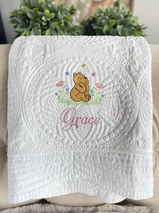 White quilt with intricate quilting patterns featuring an embroidered Classic Pooh Bear sitting in a meadow with butterflies and flowers, personalized with the name Grace, part of the Personalized Classic Pooh Bear Meadow & Butterfly Baby Quilt | Custom Name Heirloom Blanket collection.