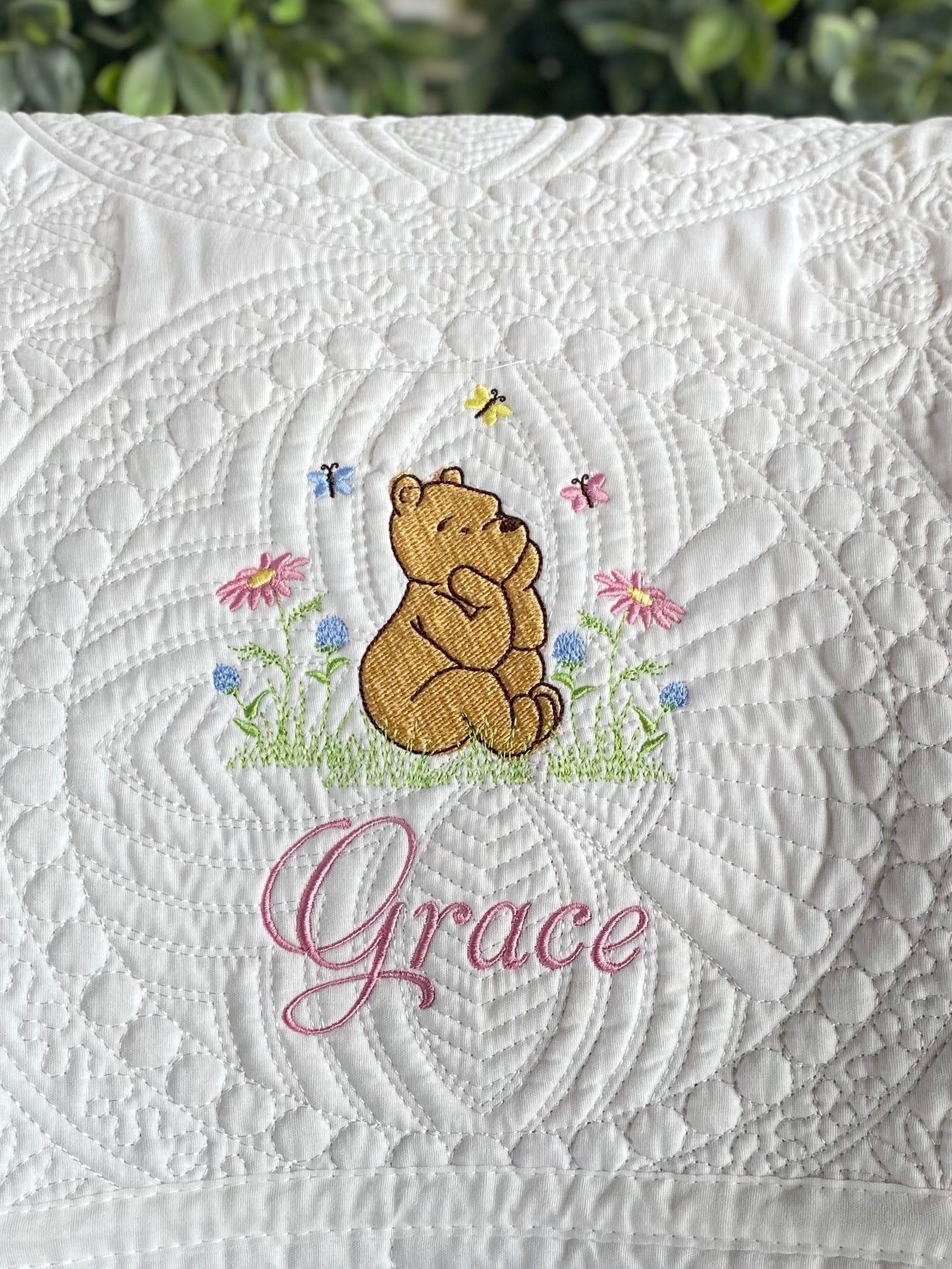 White quilt featuring an embroidered Classic Pooh Bear surrounded by colorful butterflies and flowers, with the custom name Grace stitched below on the Personalized Classic Pooh Bear Meadow & Butterfly Baby Quilt | Custom Name Heirloom Blanket.