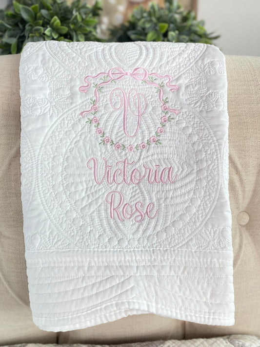 Personalized Bow & Floral Wreath Monogram Baby Quilt | Custom Name Heirloom Keepsake for Girls featuring delicate pink embroidery with a monogram and floral wreath on a white quilted background.