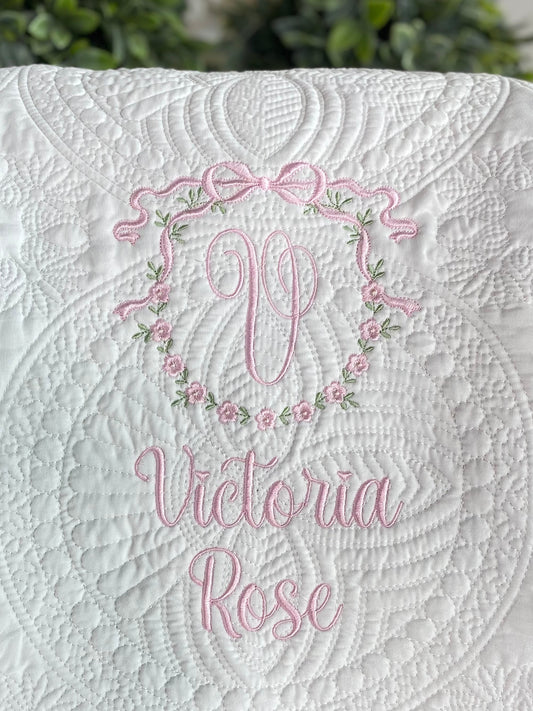 Close-up of a white quilt featuring delicate pink and green embroidered floral wreath with monogram and the name Victoria Rose, showcasing the Personalized Bow & Floral Wreath Monogram Baby Quilt | Custom Name Heirloom Keepsake for Girls.