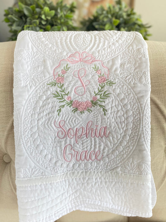 White quilt featuring intricate quilting and a personalized pink embroidered rose crest monogram with the name Sophia Grace, for the Personalized Embroidered Rose Crest Monogram Baby Quilt | Heirloom Keepsake for Girls.