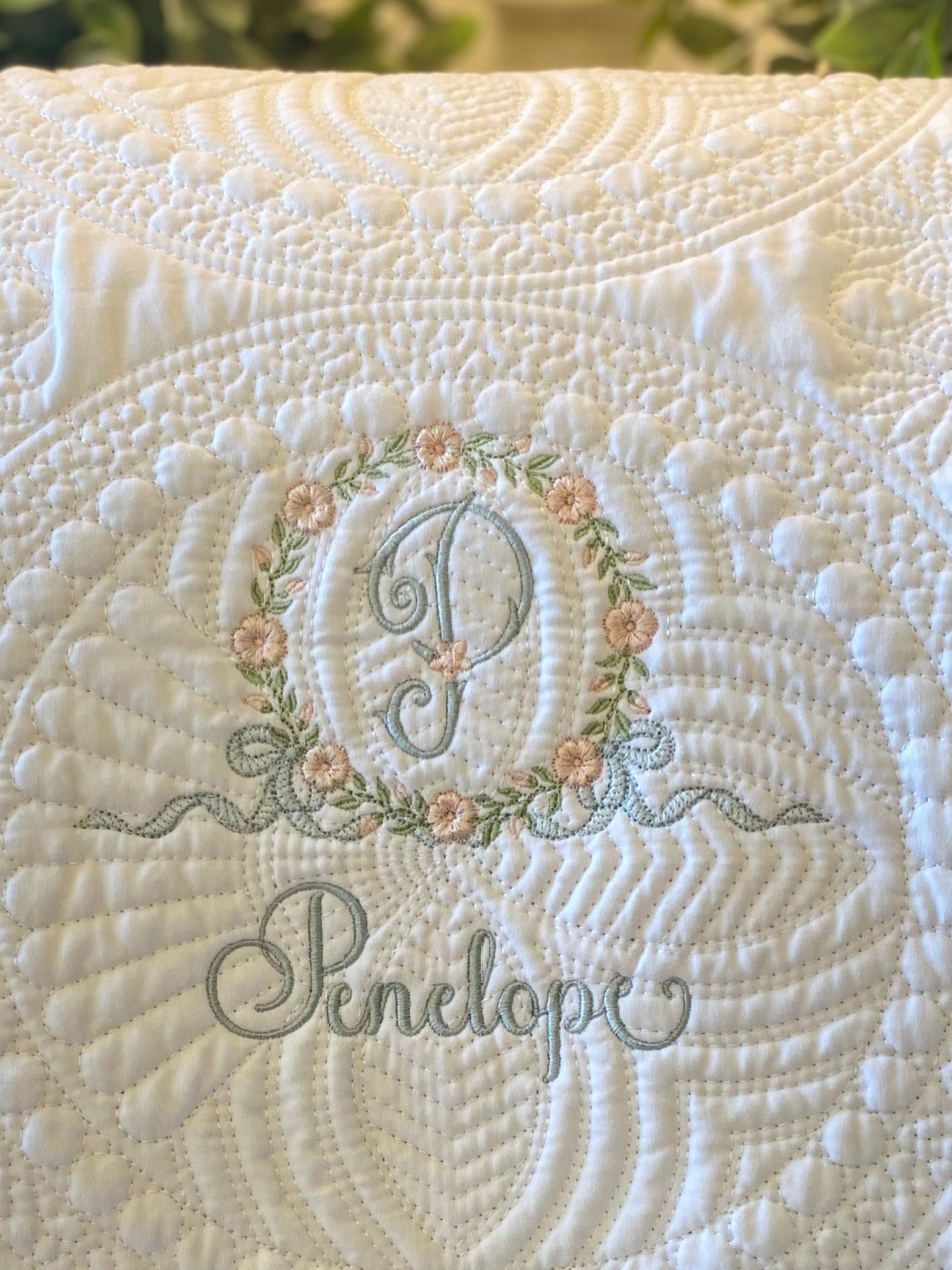 Close-up of a Personalized Embroidered Floral Wreath Monogram Baby Quilt | Heirloom Keepsake featuring elegant floral embroidery in soft colors with a monogram "P" and name "Penelope" on a white quilted fabric background.