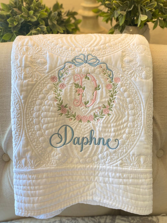 White quilt featuring intricate quilted patterns with a personalized embroidered bow and floral wreath monogram above the name Daphne, showcasing the Personalized Embroidered Bow & Floral Wreath Monogram Baby Quilt | Heirloom Keepsake design.