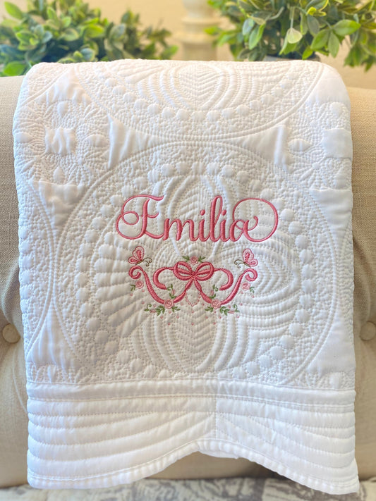 White quilt with intricate embossed floral and circular patterns, featuring personalized pink embroidery of the name Emilia with rose and butterfly bow design on the Personalized Embroidered Rose & Butterfly Bow Baby Quilt | Heirloom Keepsake.