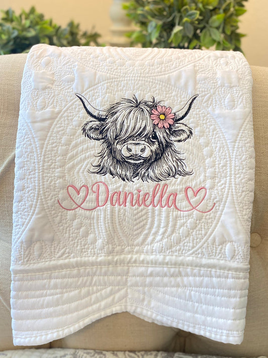 Personalized Embroidered Highland Cow Daisy Baby Quilt | Custom Name Heirloom Blanket featuring a detailed embroidered highland cow with a pink daisy and custom name Daniella on a white textured quilted fabric.