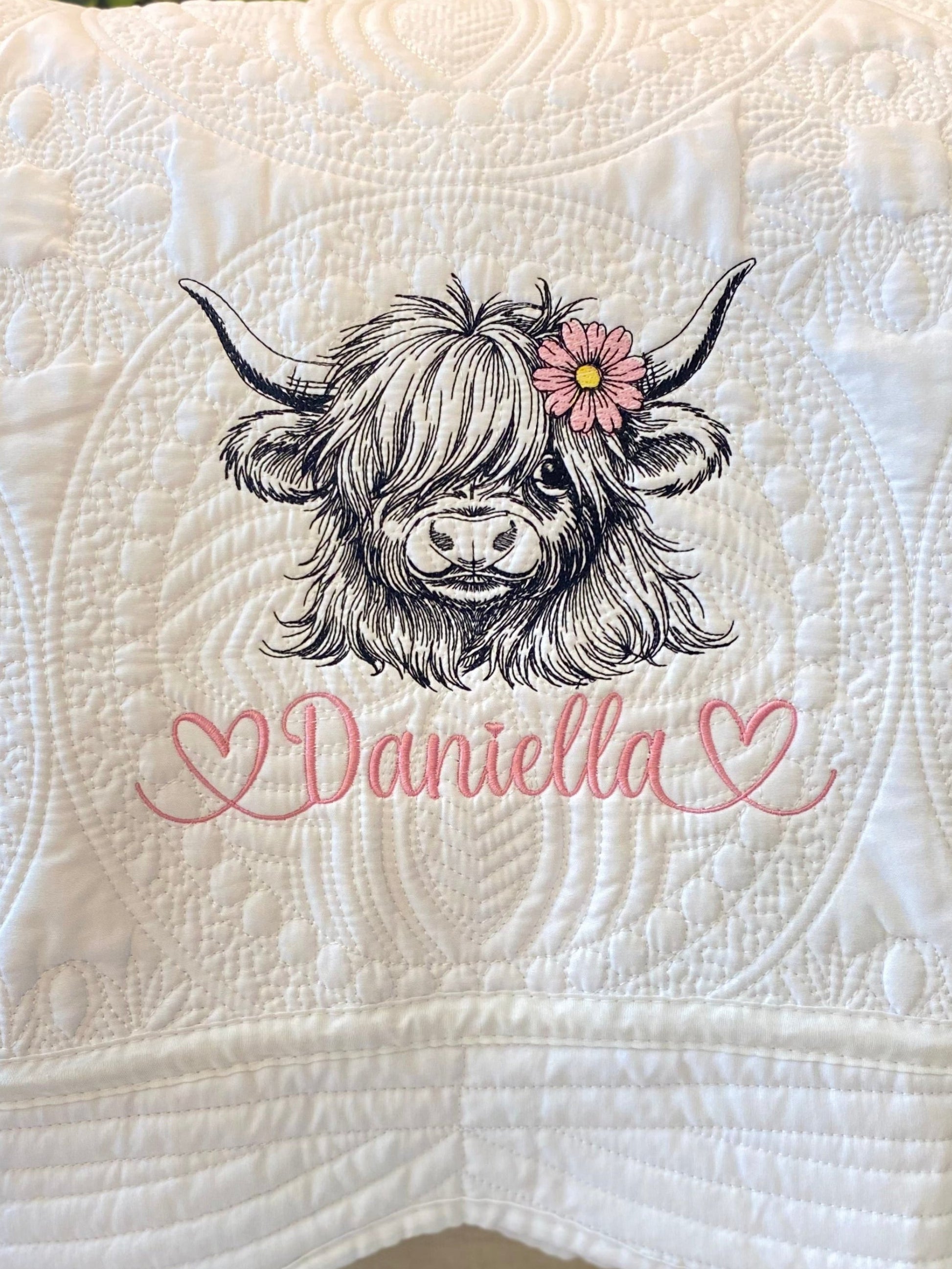 Close-up of a personalized embroidered highland cow with a pink daisy and the name Daniella in pink script on a white quilt featuring detailed stitching, showcasing the Personalized Embroidered Highland Cow Daisy Baby Quilt | Custom Name Heirloom Blanket.