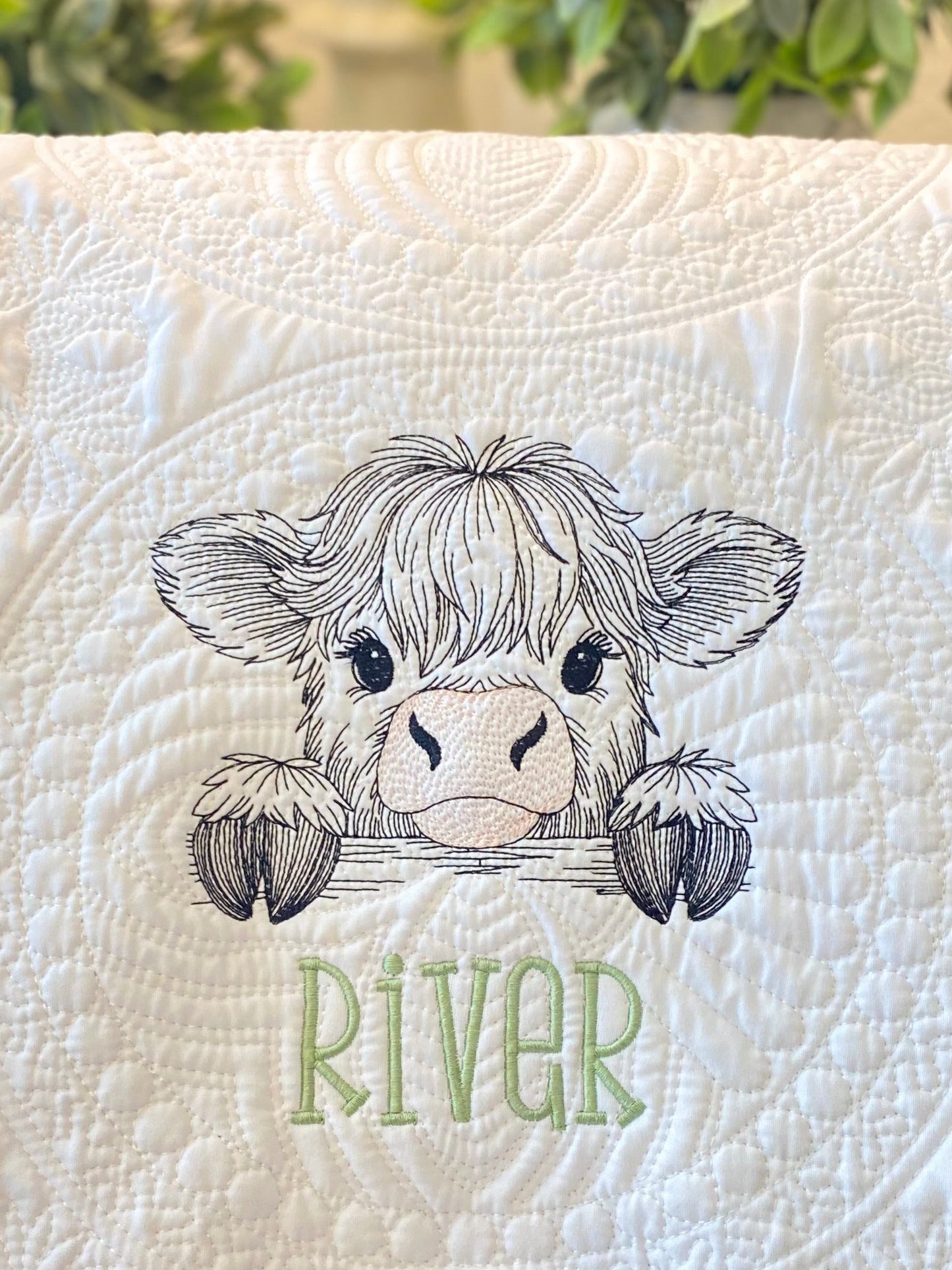 Close-up of an embroidered peeking highland cow with delicate stitching on a quilted white fabric, personalized with the name River on the Personalized Embroidered Peeking Highland Cow Baby Quilt | Custom Name Heirloom Blanket.