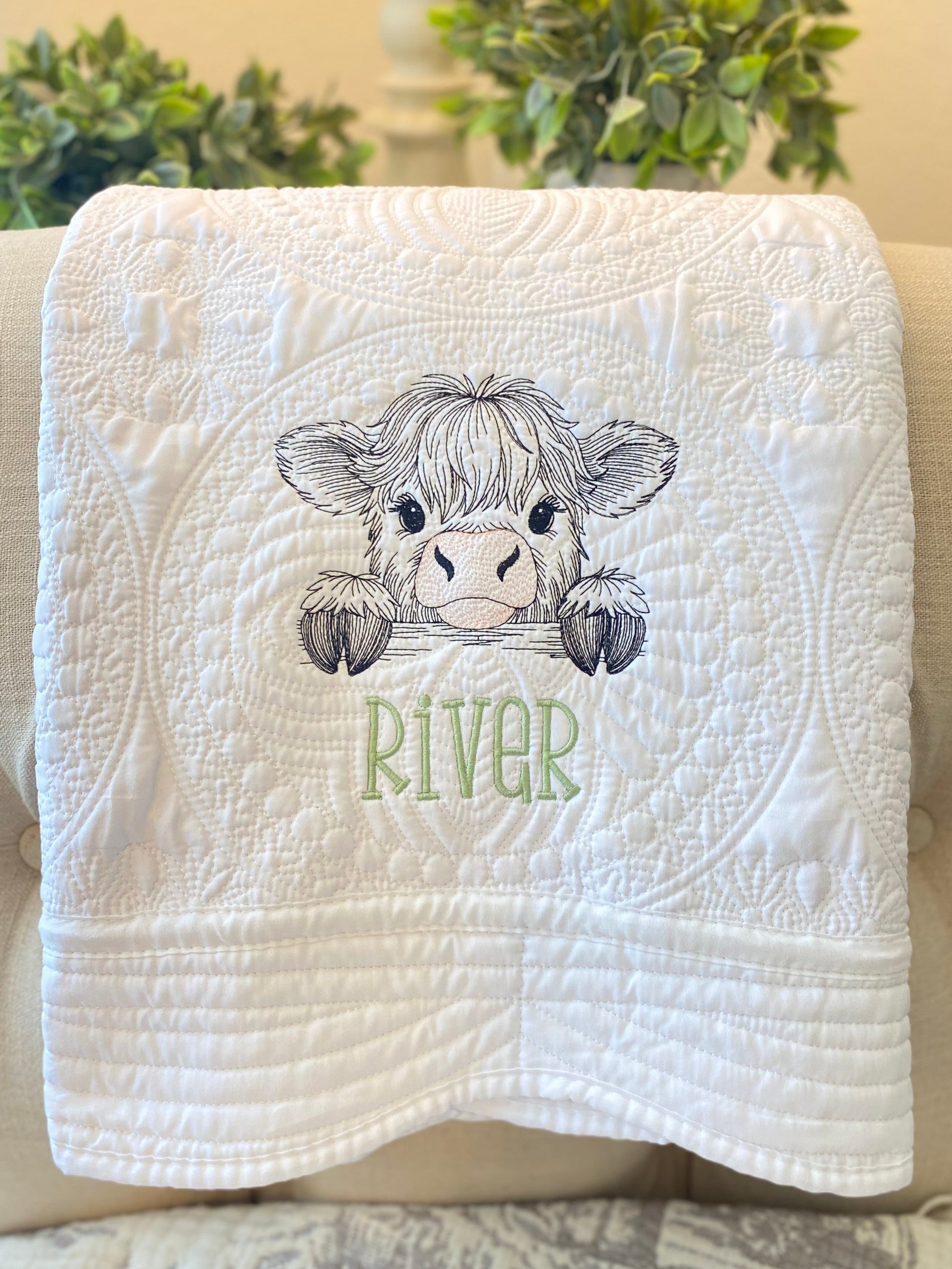 White quilt with intricate stitching featuring an embroidered peeking Highland cow and the name River in green, showcasing the Personalized Embroidered Peeking Highland Cow Baby Quilt | Custom Name Heirloom Blanket.