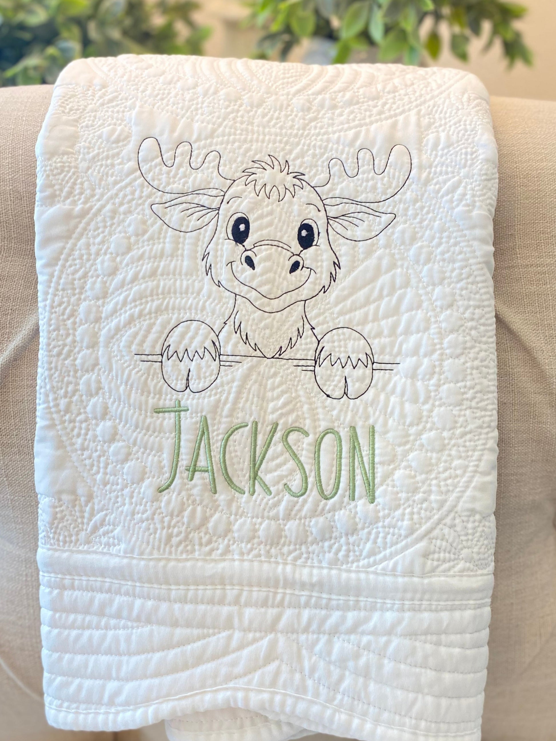 White quilt featuring an embroidered moose design and a custom name in green, showcasing the Personalized Embroidered Moose Baby Quilt | Custom Name Heirloom Blanket as a handmade heirloom baby gift.