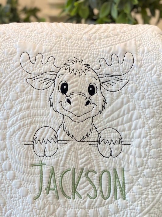 Close-up of a cream-colored Personalized Embroidered Moose Baby Quilt | Custom Name Heirloom Blanket featuring a smiling moose outline stitched above a custom name in green thread on a textured quilted background.