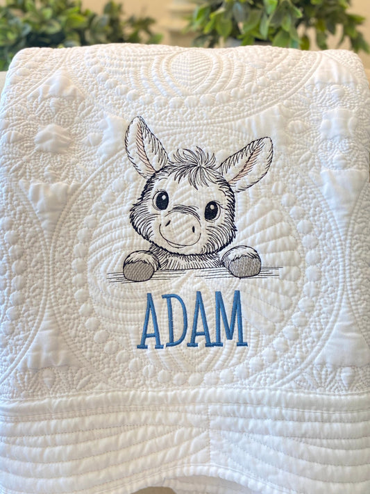 Close-up of a white quilt featuring intricate embroidery with a cute donkey design and the name Adam, showcasing the personalized detail on the Personalized Embroidered Donkey Baby Quilt | Custom Name Heirloom Blanket.