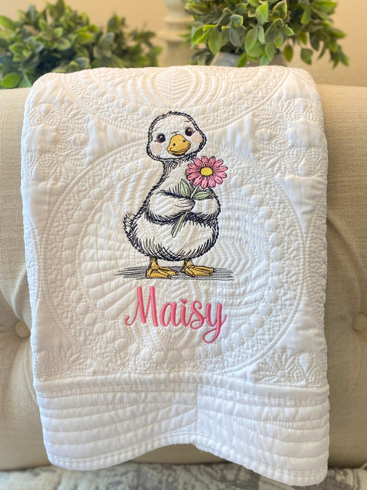 White quilt featuring intricate stitching with an embroidered duck holding a pink flower and the name Maisy in pink script, part of the Personalized Embroidered Duck with Flower Baby Quilt | Custom Name Heirloom Blanket collection.