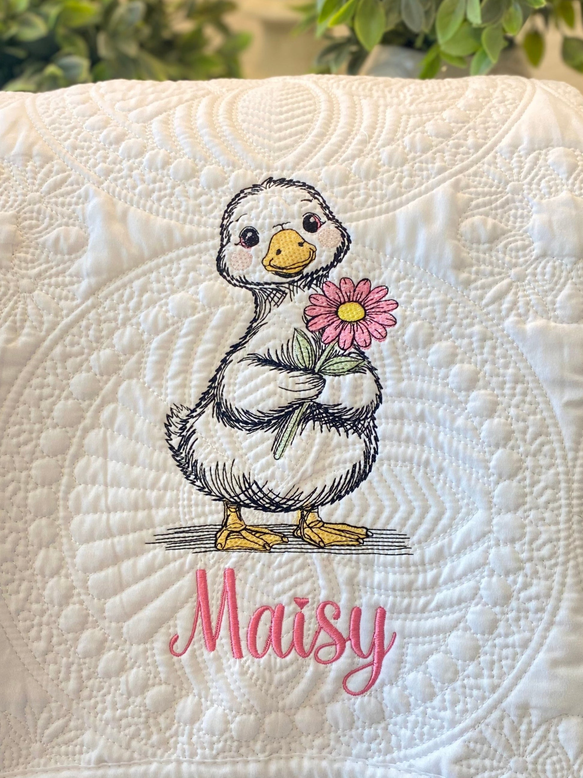 Close-up of a Personalized Embroidered Duck with Flower Baby Quilt | Custom Name Heirloom Blanket featuring a cute duck holding a pink flower with the name Maisy embroidered below on a textured white quilted background.