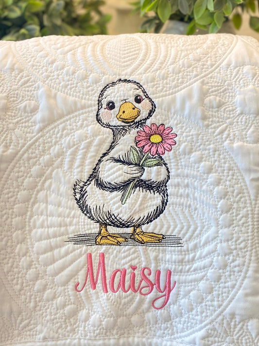 Close-up of a Personalized Embroidered Duck with Flower Baby Quilt | Custom Name Heirloom Blanket featuring a cute duck holding a pink flower with the name Maisy embroidered below on a textured white quilted background.