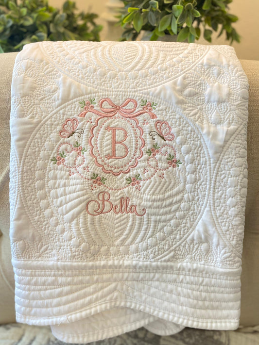 Soft white quilt with intricate scalloped stitching featuring an embroidered pink bow, butterflies, floral details, and a personalized monogram "B" with the name Bella, showcasing the Personalized Embroidered Bow & Butterfly Scallop Monogram Baby Quilt | Heirloom Keepsake.