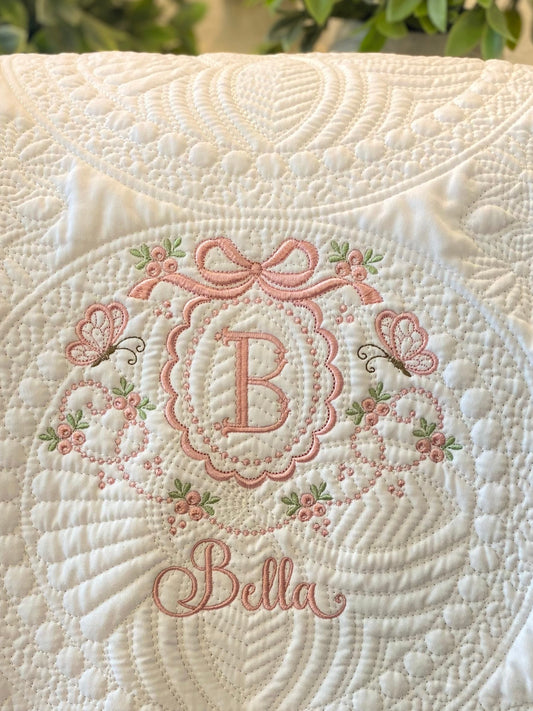 Close-up of a soft white quilt featuring delicate scallop quilting and an embroidered pink bow, butterflies, floral details, and the monogram letter B with the name Bella on the Personalized Embroidered Bow & Butterfly Scallop Monogram Baby Quilt | Heirloom Keepsake.