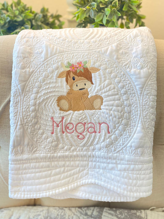 Personalized Embroidered Highland Cow Flower Crown Baby Quilt | Custom Name Heirloom Blanket with a cute embroidered brown highland cow wearing a flower crown and the name Megan stitched below on a white quilted background.