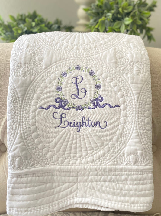 Soft white quilt featuring intricate circular quilting patterns and a delicate embroidered floral wreath monogram with the letter L and the name Leighton in lavender, showcasing the Personalized Embroidered Floral Wreath Monogram Baby Quilt | Heirloom Keepsake.