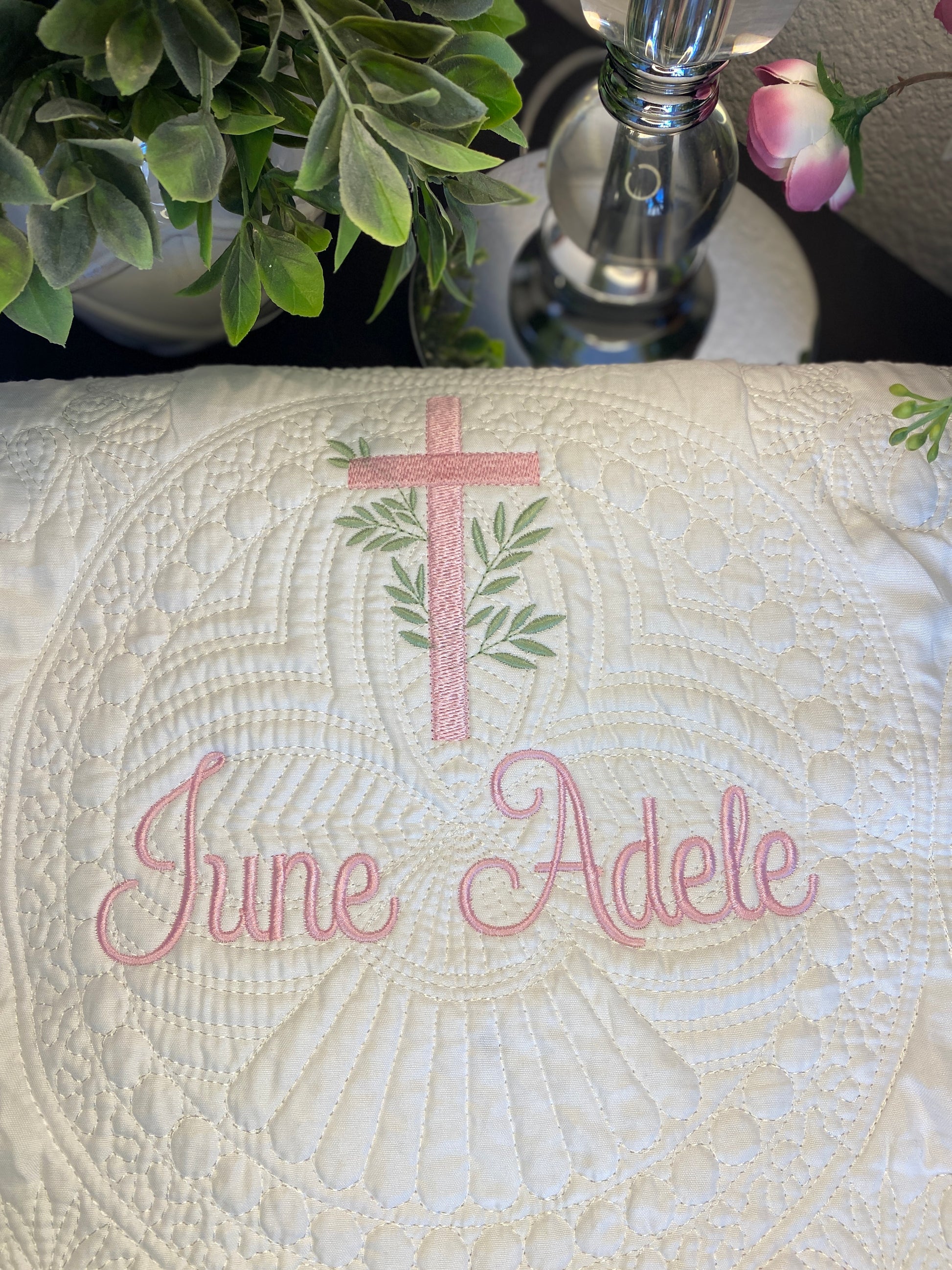 Close-up of a white quilted fabric with delicate embroidered details including a pink cross with laurel branches and the name June Adele in pink script, part of the Personalized Laurel Branch Cross Baptism Gift Set | Custom Quilt Bib & Burp Cloth Christening Heirloom.