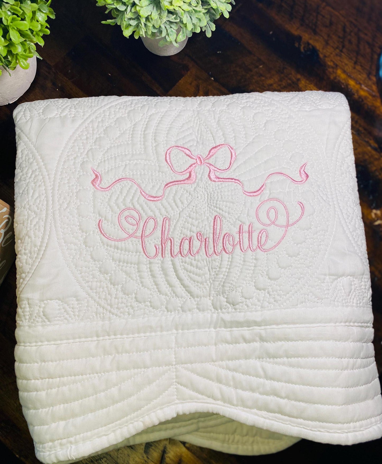 Personalized Pastel Pink Ribbon Bow Baby Quilt | Custom Name Heirloom Blanket for Girls with delicate pink embroidery of a ribbon bow and the name Charlotte on a white quilted fabric.