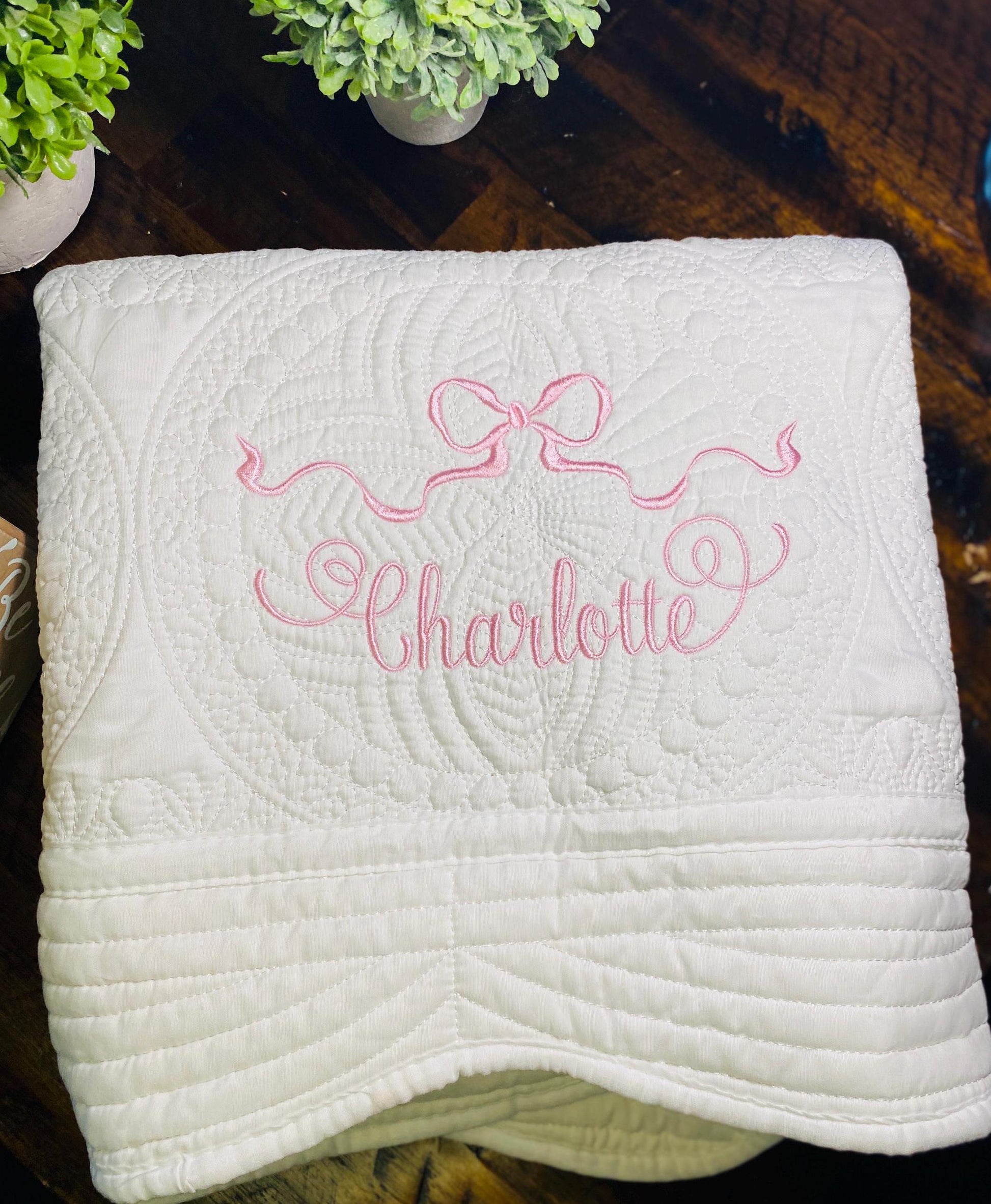 Personalized Pastel Pink Ribbon Bow Baby Quilt | Custom Name Heirloom Blanket for Girls with delicate pink embroidery of a ribbon bow and the name Charlotte on a white quilted fabric.