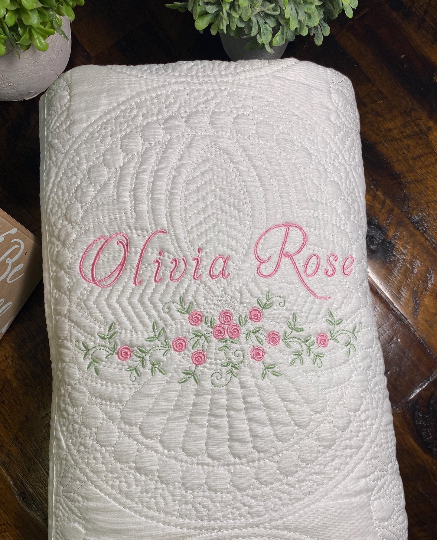 White quilt with intricate stitching and delicate pink floral embroidery, personalized with the name Olivia Rose, showcasing the Personalized Delicate Floral Rose Swag Baby Quilt | Custom Embroidered Heirloom Blanket.
