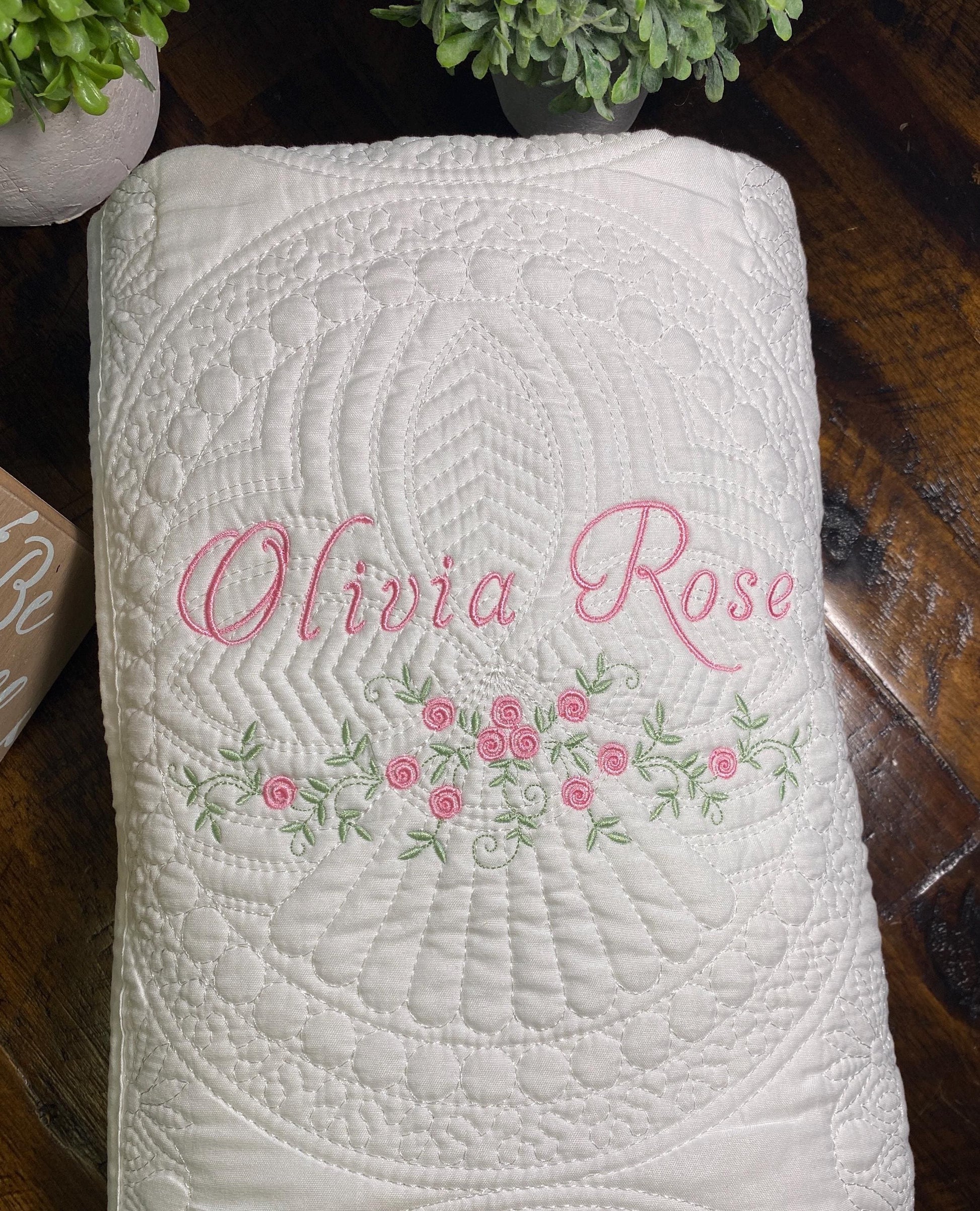 White quilt with intricate stitching and delicate pink floral embroidery, personalized with the name Olivia Rose, showcasing the Personalized Delicate Floral Rose Swag Baby Quilt | Custom Embroidered Heirloom Blanket.